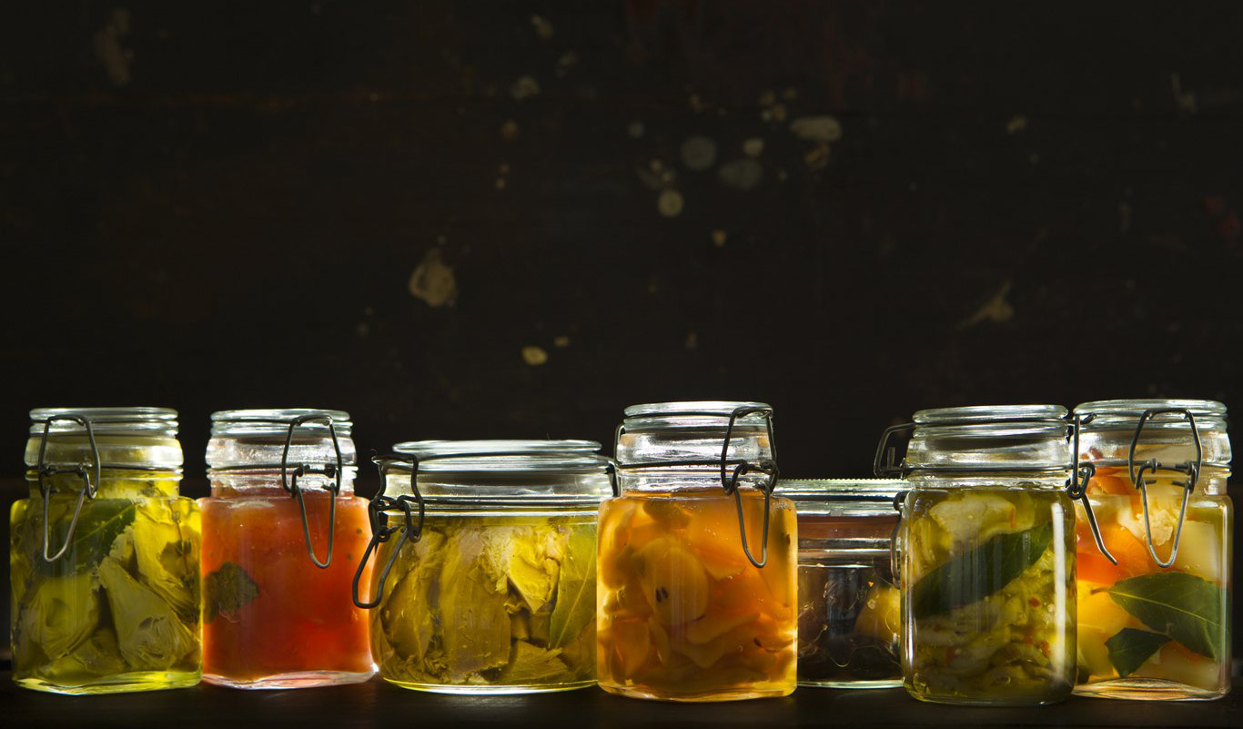 A variety of preserved food in glass jar. From left to right: artichokes, tomatoes, tuna, pears, olives, eggplants, mixed vegetables.