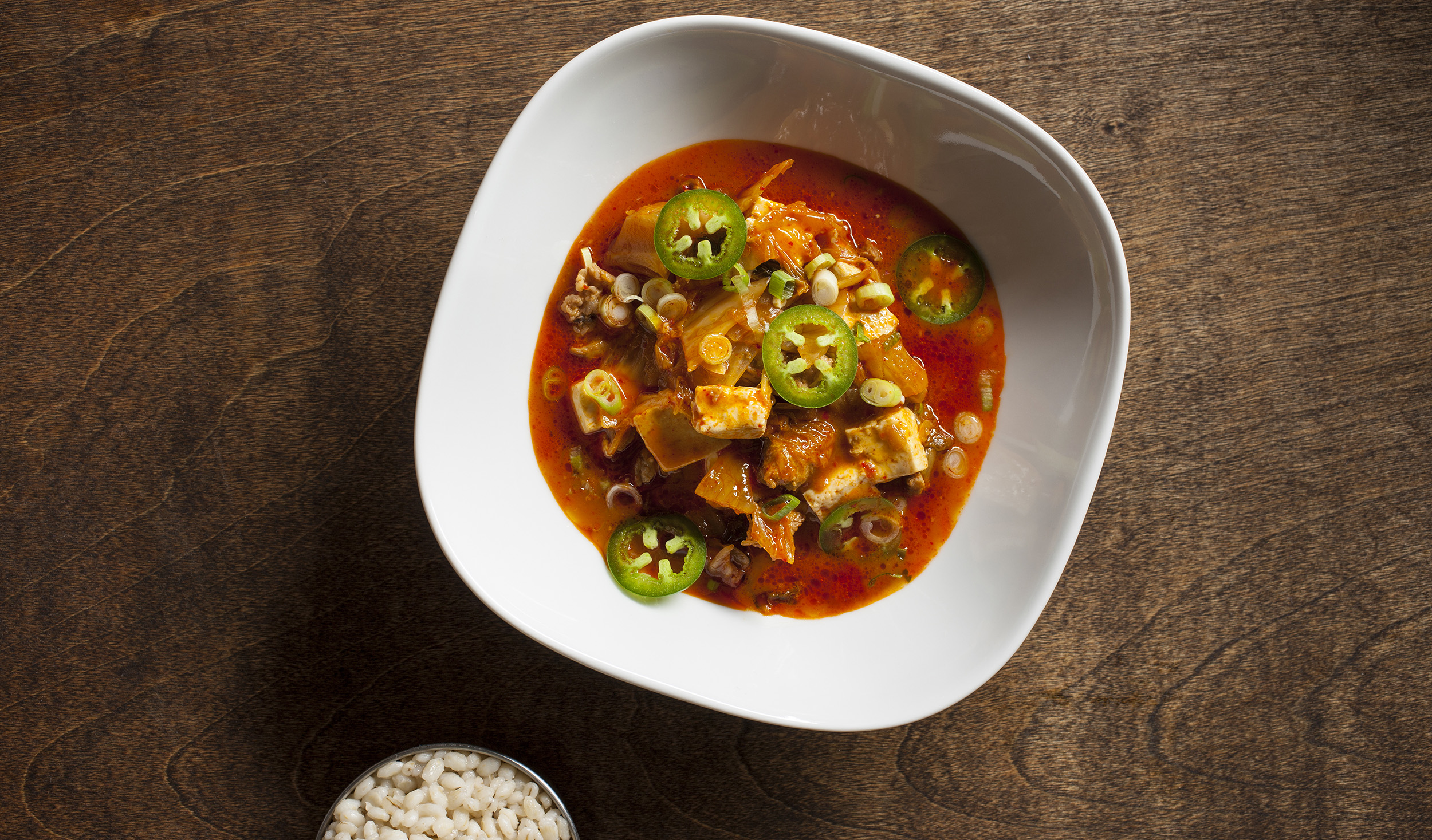 Kimchi stew with tofu and pork garnished with jalapeno served with a side of barley.