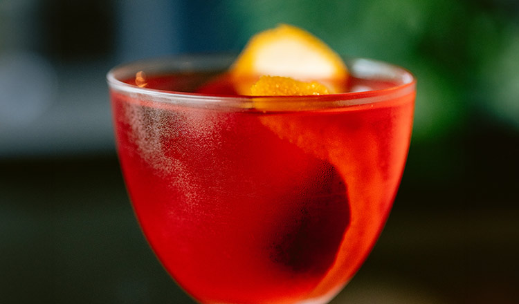 Image of a red colored amaro cocktail with an orange peel.