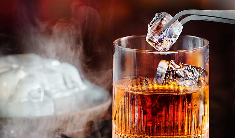 Image of a glass of bourbon with ice being placed in it with tongs.