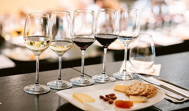 Image of five glasses of wine on a table with a plate of cheeses, nuts, fruits, and crackers.