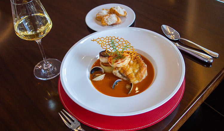 Image of a seafood dish with fish, scallops, shrimp, sauce, and garnish, served with wine and bread on a table.