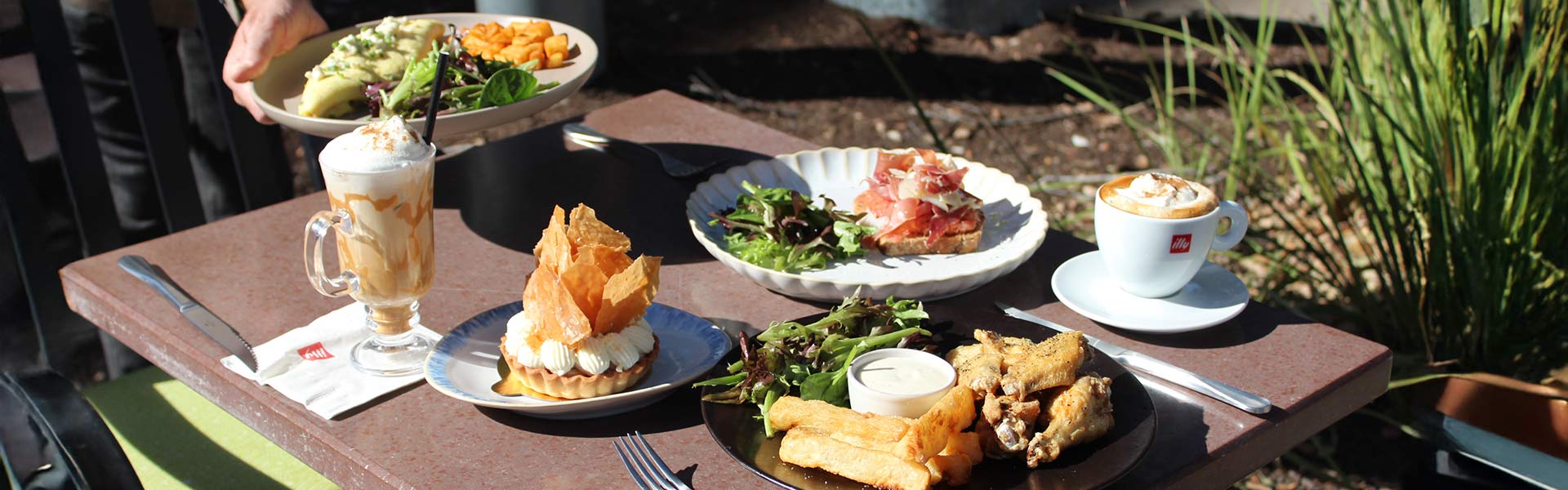 CIA Bakery Café outdoor table with coffee drinks, pastries, salads, and sandwiches served in sunlight on a patio surrounded by greenery.