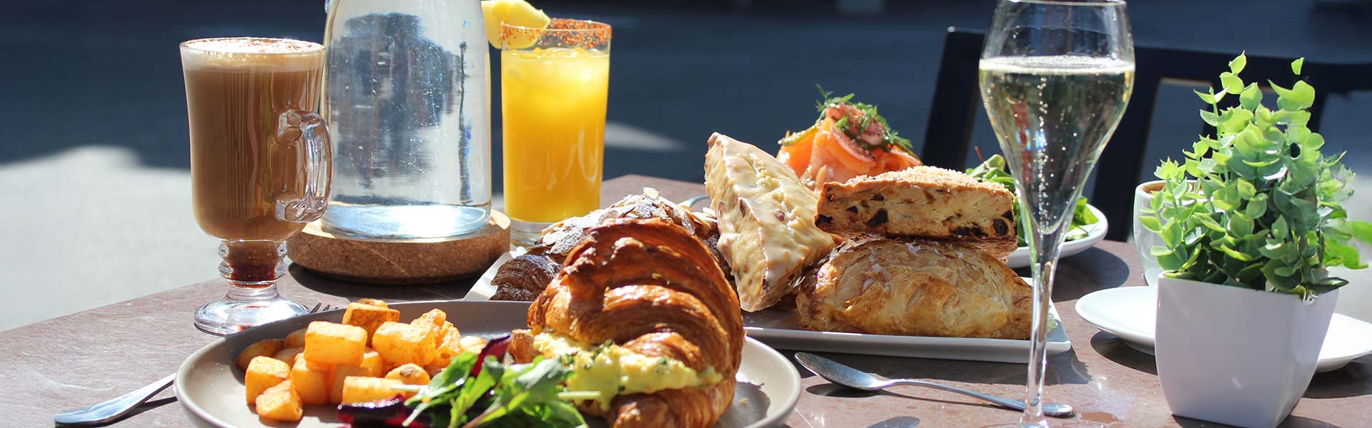 CIA Bakery Café outdoor table is set for brunch with croissants, pastries, potato cubes, a latte, orange juice, and champagne in sunlight.
