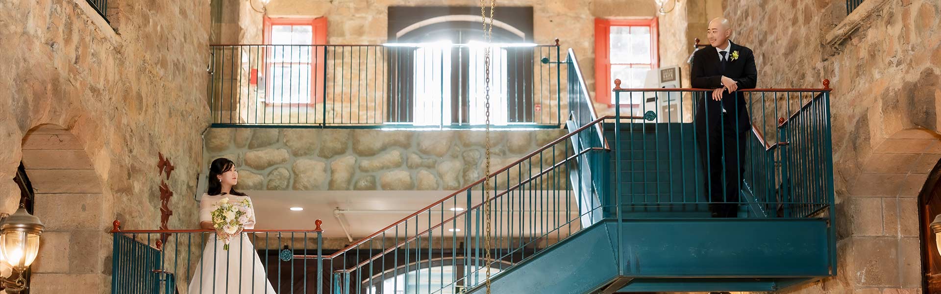 A bride stands on a stairway, looking towards the groom on a balcony above at CIA at Greystone.