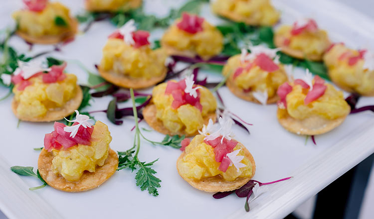 White platter of crackers topped with yellow mash, pink garnish, and edible flowers on greens.